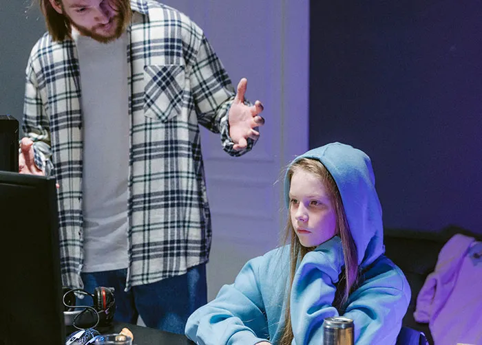 A man stands over a girl in a blue hoodie who is looking at a computer, depicting unsettling sentences in their interaction.