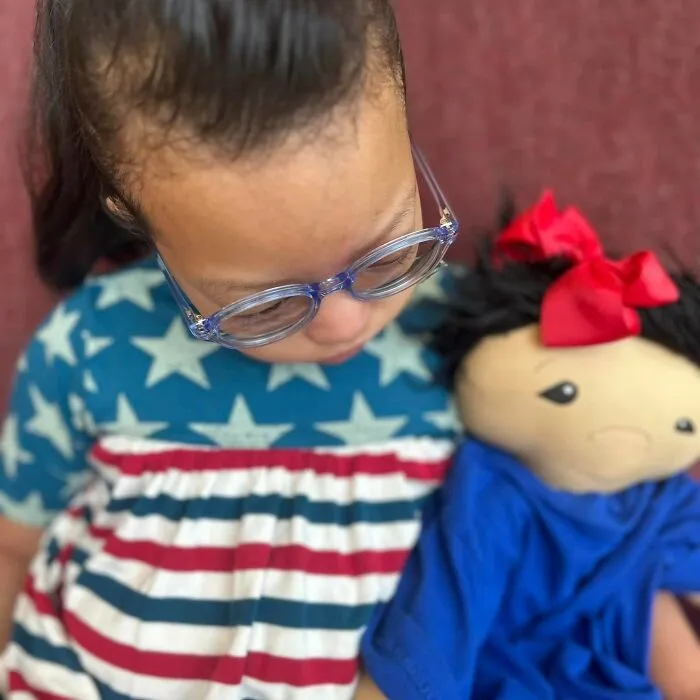 A young girl with a rare health condition wearing glasses looks down at a doll tailored to her needs.