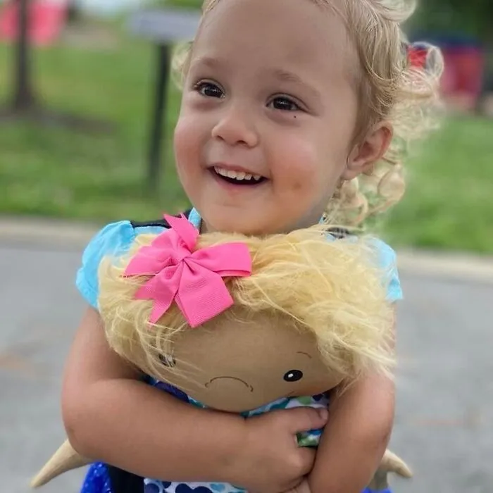 A happy toddler with blonde curly hair hugs a doll with blonde hair, a doll that fits children's needs.