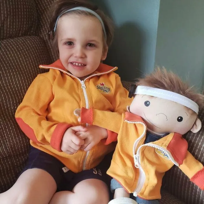 A smiling child with a rare health condition sits beside a doll designed to fit their specific needs.