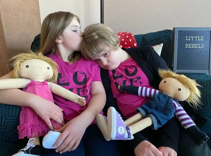 Two girls with rare health conditions hug their dolls, representing their unique needs.