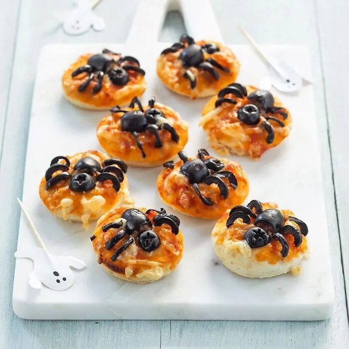 Mini Halloween snacks pizza topped with olive spiders arranged on a white cutting board for spooky treats.
