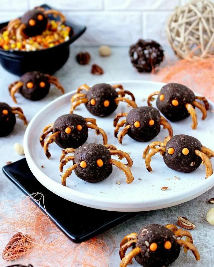 Halloween snacks shaped like spiders with pretzel legs and orange candy eyes displayed on a white plate.