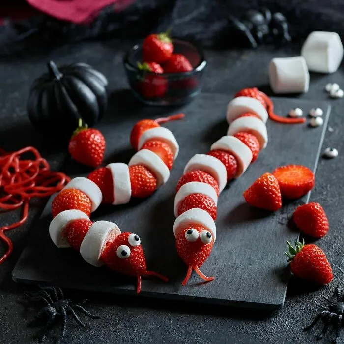 Halloween snacks shaped like snakes with strawberries, marshmallows, and candy eyes on a black serving board.