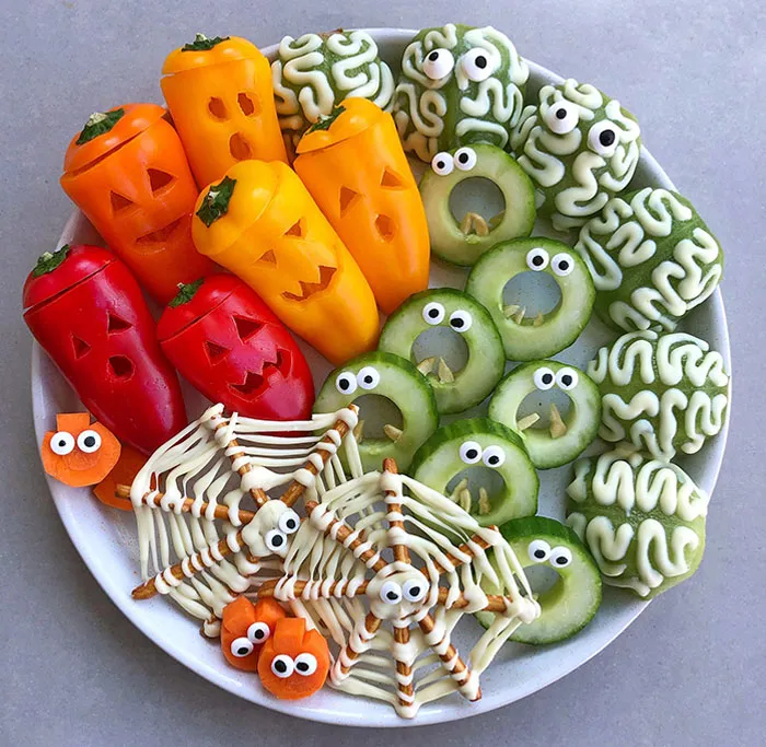 Plate of Halloween snacks including carved mini peppers, cucumber slices with eyes, brain-shaped snacks, and spider web pretzels with eyes.