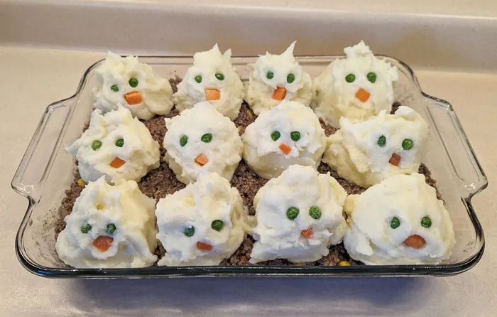 Halloween snacks made with mashed potatoes and ground beef, shaped like ghost faces with peas and carrots as eyes and mouths.