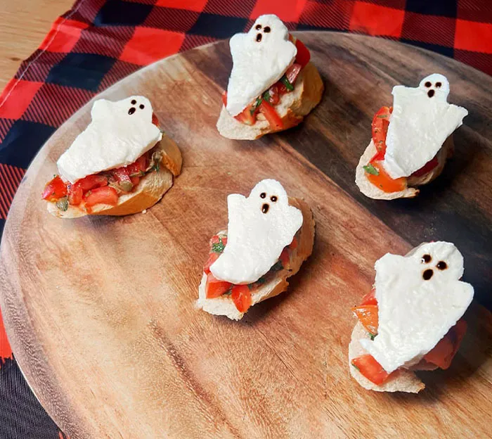 Halloween snacks featuring ghost-shaped toppings on bread with tomato and herbs, arranged on a wooden serving board.