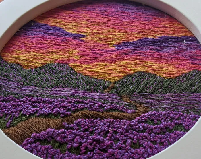 Colorful embroidery design of a vibrant landscape with purple flowers and a sunset sky in an embroidery hoop.