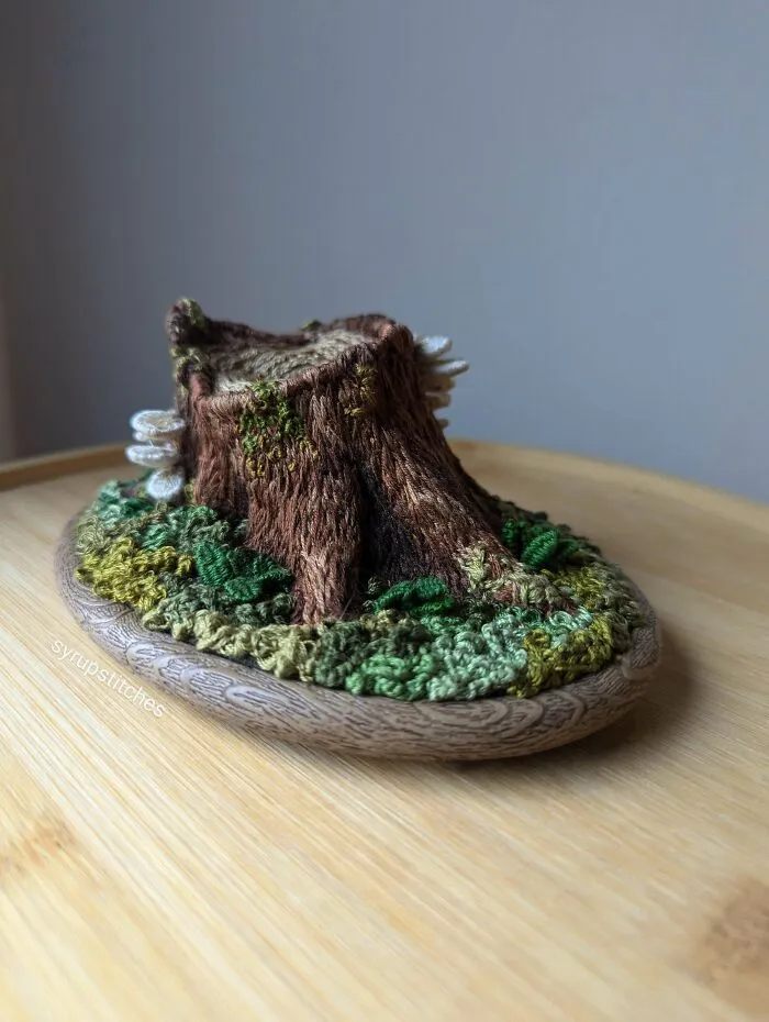 Intricate embroidery design of a tree stump with green foliage on a wooden surface, showcasing beautiful embroidery designs.