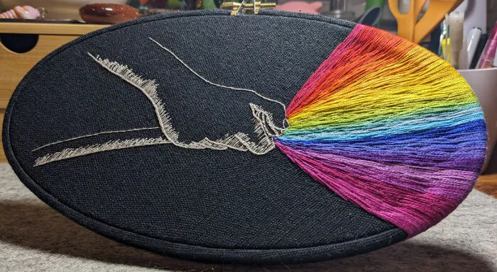 Hand embroidery design of a hand pulling colorful rainbow threads on black fabric, showcasing beautiful embroidery art.