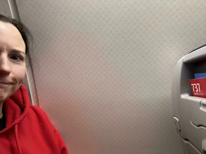 Person in a red hoodie with an irritated expression next to a wall on an airplane.