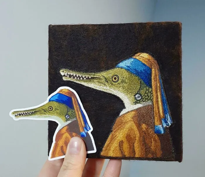 Hand holding embroidered design of a crocodile with blue and orange clothing, showcasing beautiful embroidery designs.