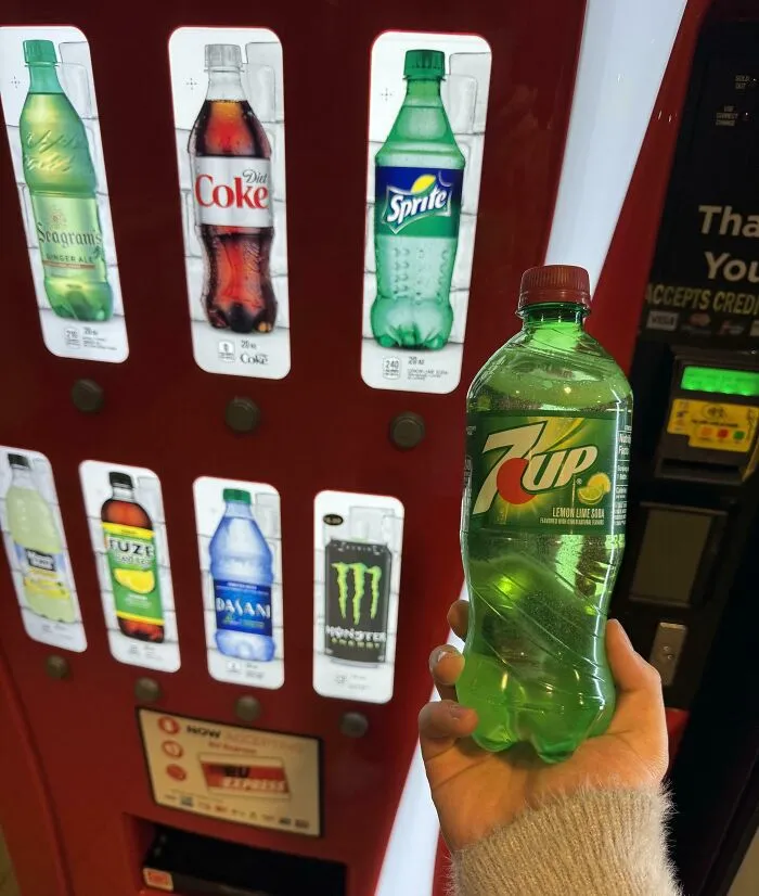 Hand holding a 7UP bottle in front of a vending machine display.