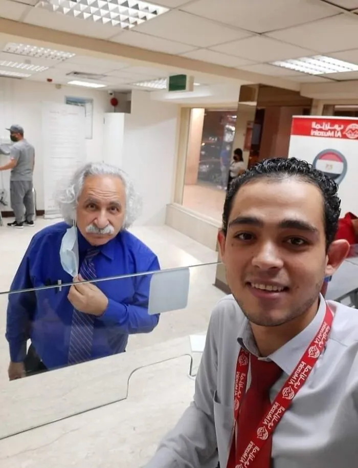 Man in blue shirt resembles famous figure, standing near another man in red tie, behind a glass partition, showcasing uncanny doppelgangers.