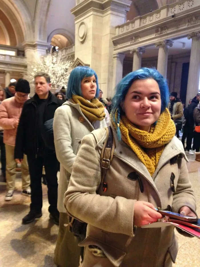 Two women with blue hair and yellow scarves, resembling doppelgangers, standing in a crowded hall.