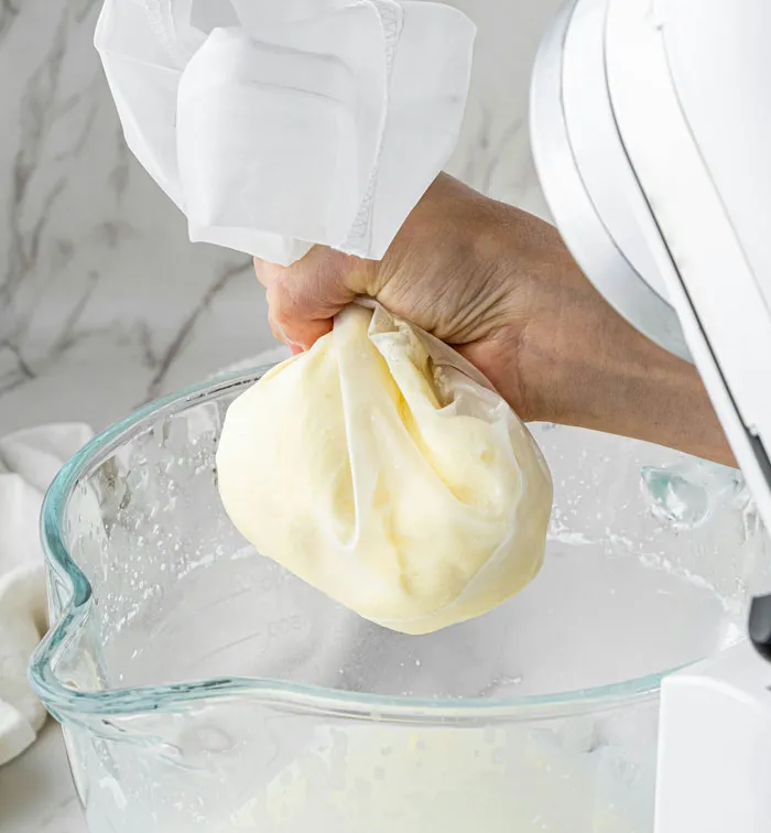 Hand squeezing homemade butter in a cloth over a glass bowl, illustrating a frugal choice.