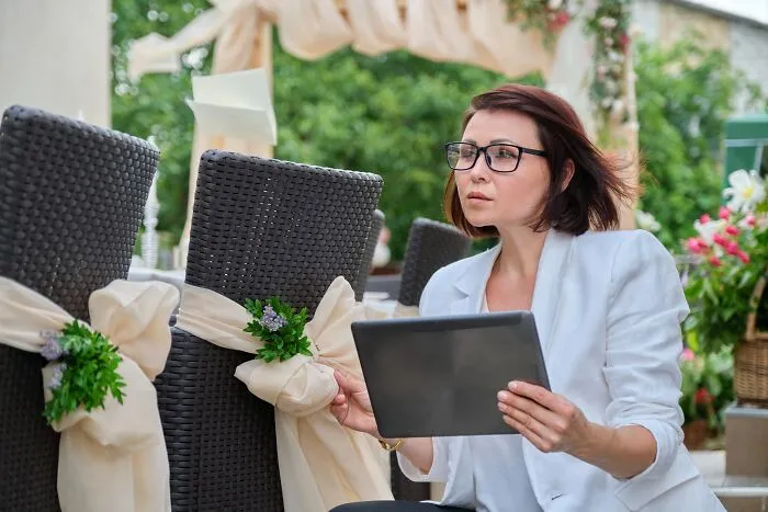 Woman in glasses holding a tablet, surrounded by wedding decorations, looking focused in an outdoor setting.