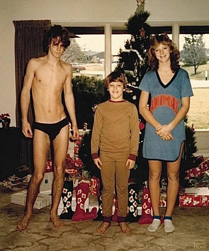 Awkward family Christmas photo with two teenagers and a kid in front of a decorated tree and presents.