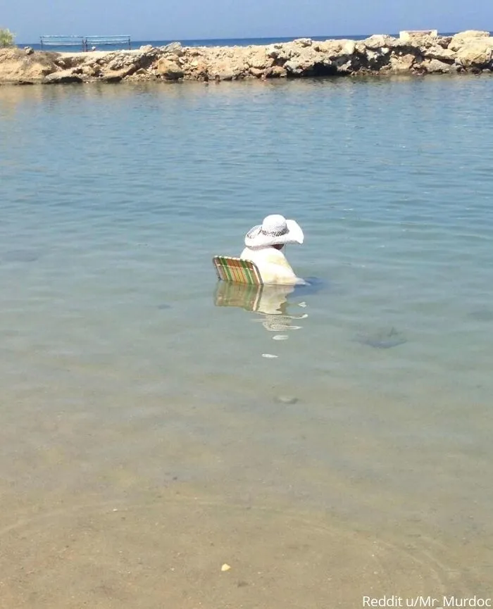 Person in beach chair submerged in shallow water, wearing a sun hat.