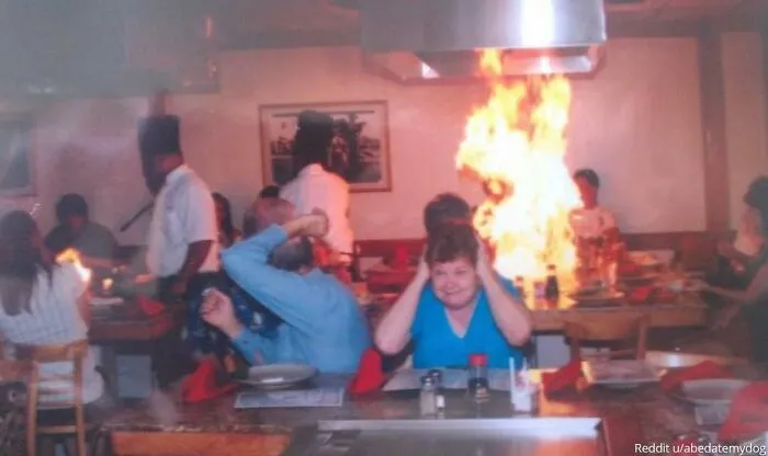 Family photo in a restaurant with a chef, fire erupting in background, and people reacting in shock, during a hibachi dinner.
