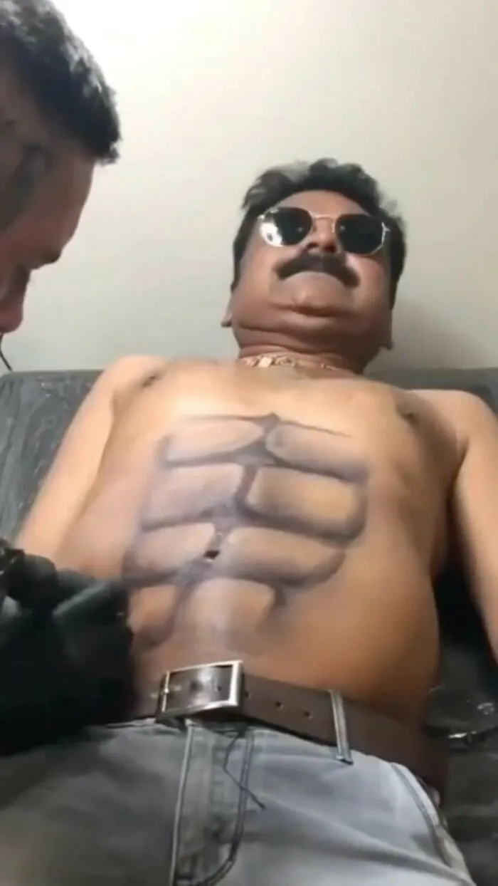 Man with a tattooed six-pack fail, lying on a tattoo chair, wearing sunglasses and a belt.