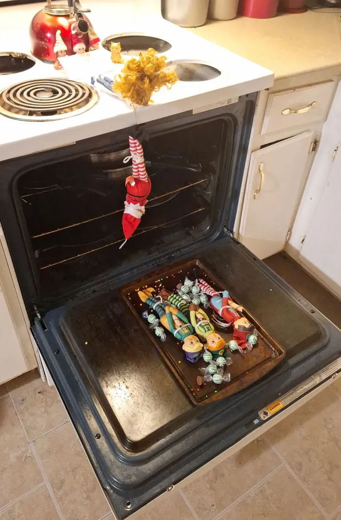 Elf On The Shelf idea with elf dangling over oven and toy elves on baking tray in a playful mischief scene.
