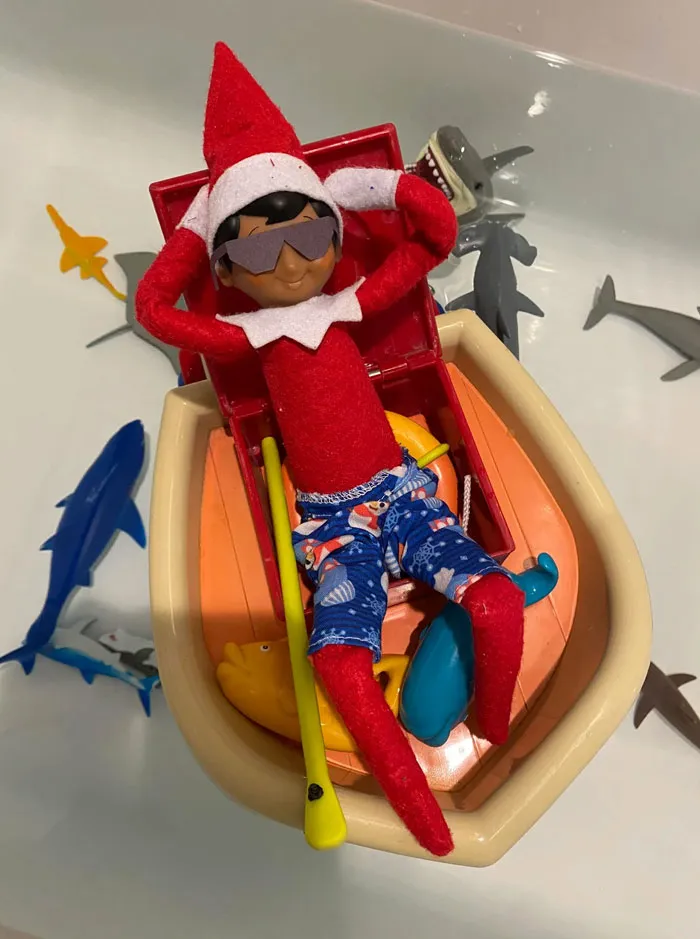 Elf on the Shelf relaxing in a toy boat with sunglasses and swim trunks surrounded by toy sharks in a bathtub.