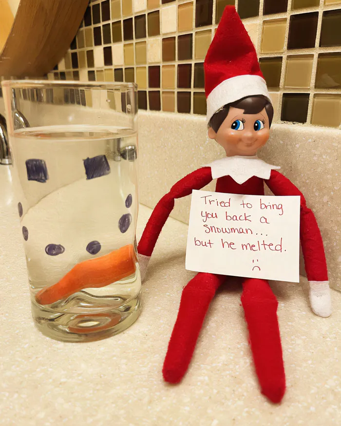 Elf On The Shelf idea with elf holding sign about melted snowman next to glass with drawn snowman features and carrot.