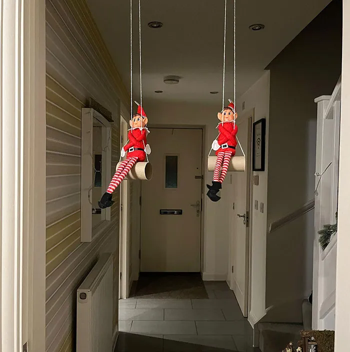 Two Elf On The Shelf dolls sitting on swings made from toilet paper rolls hanging from the ceiling in a hallway.