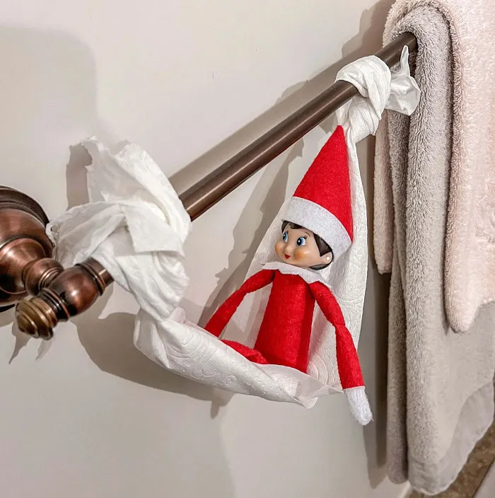 Elf On The Shelf idea with elf sitting in a toilet paper hammock hanging on a bathroom towel bar.
