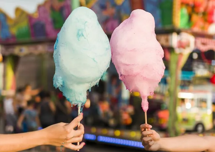 Two hands holding blue and pink cotton candy at a colorful fairground, evoking happiness.