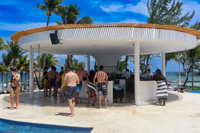 Crowded beach bar with people in swimwear, palm trees, and ocean view, representing hyped travel destinations.