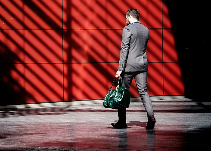 Man in a suit walking past a red wall, holding a green bag.