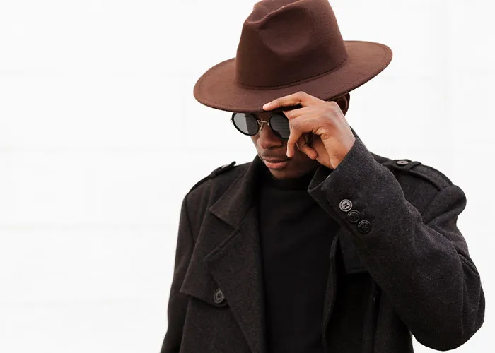 Man in a brown hat and coat, adjusting glasses; embodying a cool, mysterious vibe.