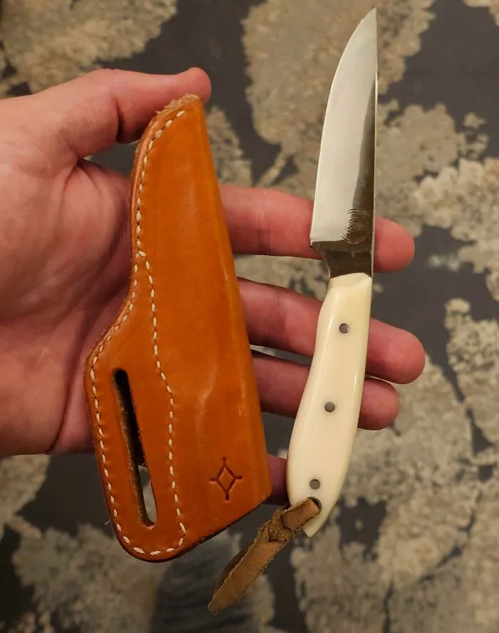 Hand holding a small knife with a white handle next to a brown leather sheath, suitable for a good-funny-bosses theme.