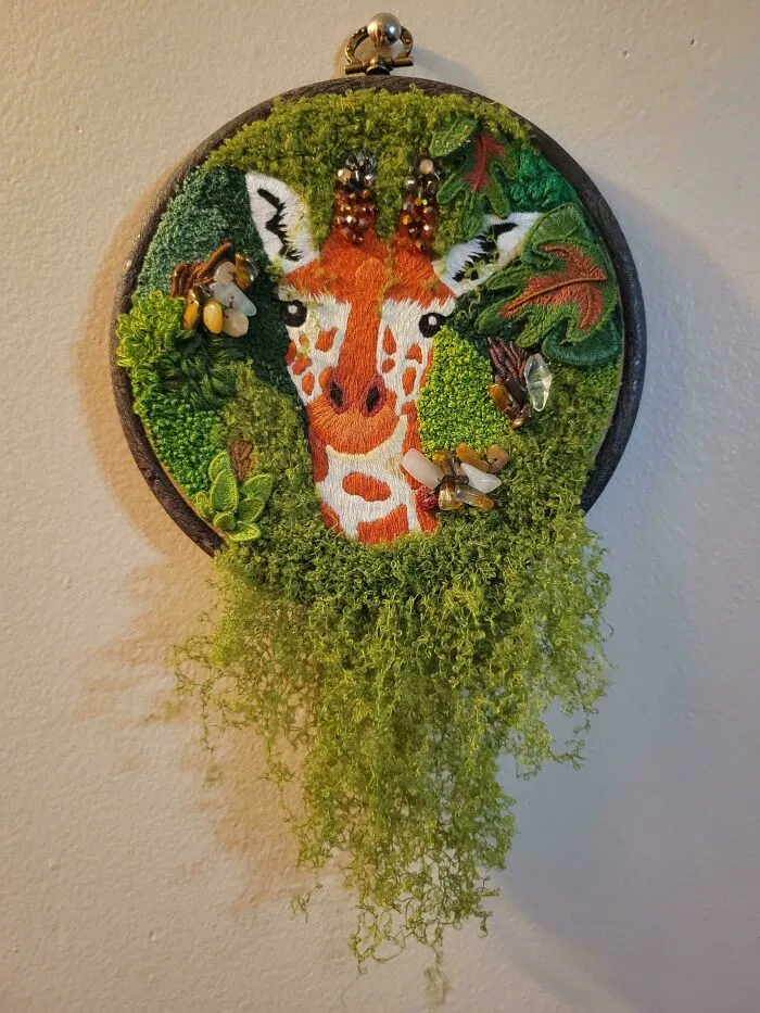 Embroidery design of a giraffe surrounded by green leaves with textured green thread and bead embellishments in a hoop.