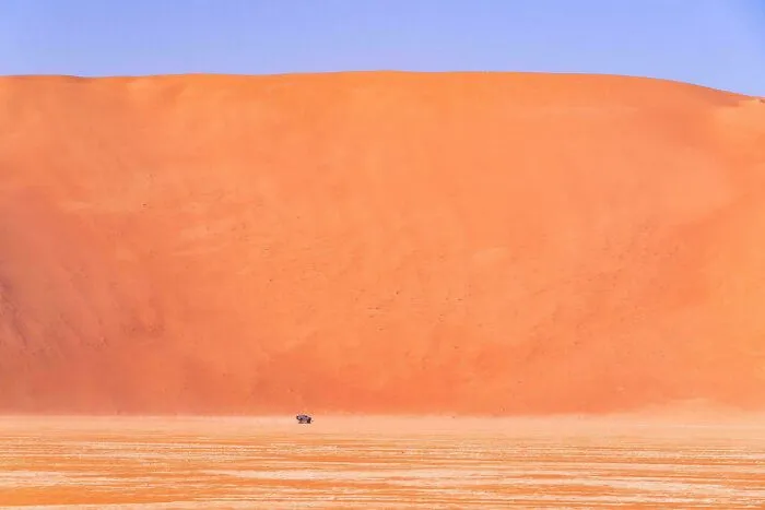 Massive orange sand dune towering over a small vehicle in a vast desert, illustrating megalophobia scale and size contrast.