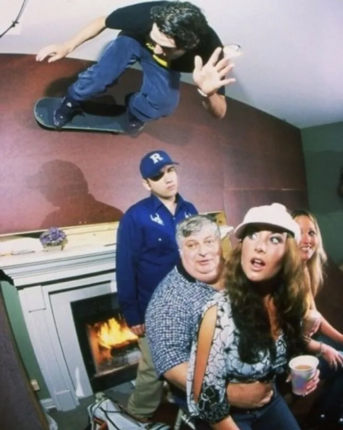 Skateboarder defying gravity indoors, over a surprised group by a fireplace, showcasing weirdness.