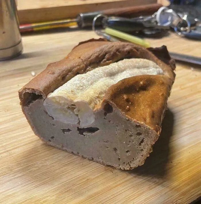 A baked dish resembling bread with a peeled banana partially embedded on top on a wooden cutting board.