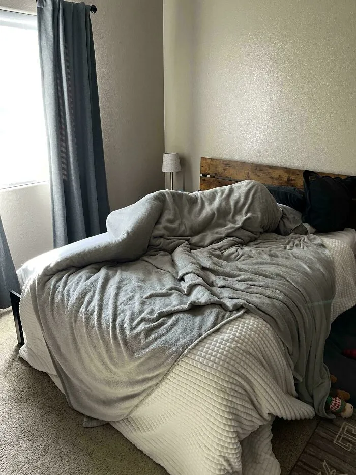 A disturbingly human-like shape under a gray blanket on an unmade bed, creating an oddly terrifying scene.