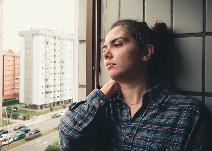 Person in plaid shirt, looking out a window with a reflective expression; urban buildings in the background.