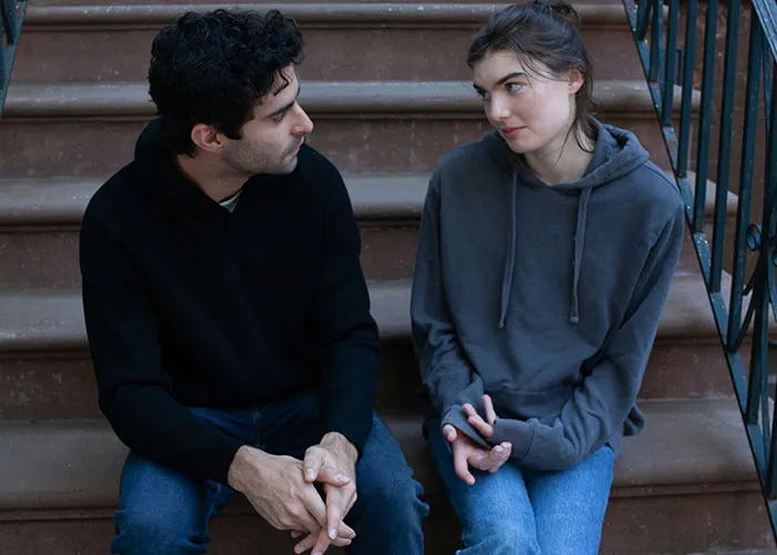 Two people sitting on steps, sharing secretive conversation, wearing casual hoodies and jeans.