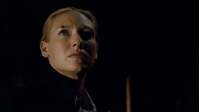 A determined woman with a scar on her forehead stands in a dimly lit setting, embodying a strong female protagonist.