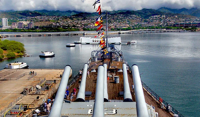 View from the deck of a large battleship with flags, stationed at a harbor with mountains in the background.