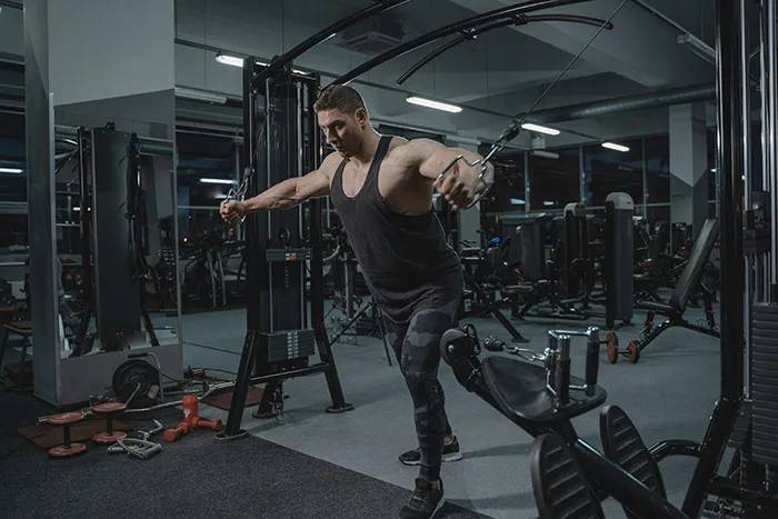 Man exercising in gym, wearing a black tank top, surrounded by fitness machines and weights, showcasing unique fitness routine.