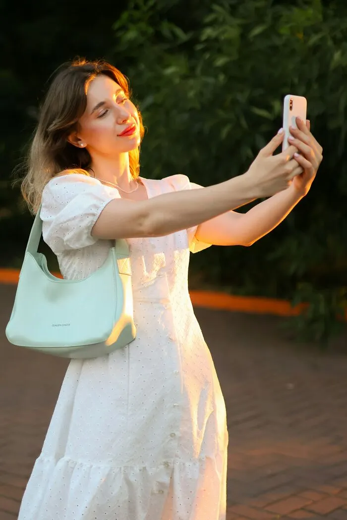 Woman in a white dress taking a selfie with a smartphone, secretly judged by others for her social media habits.