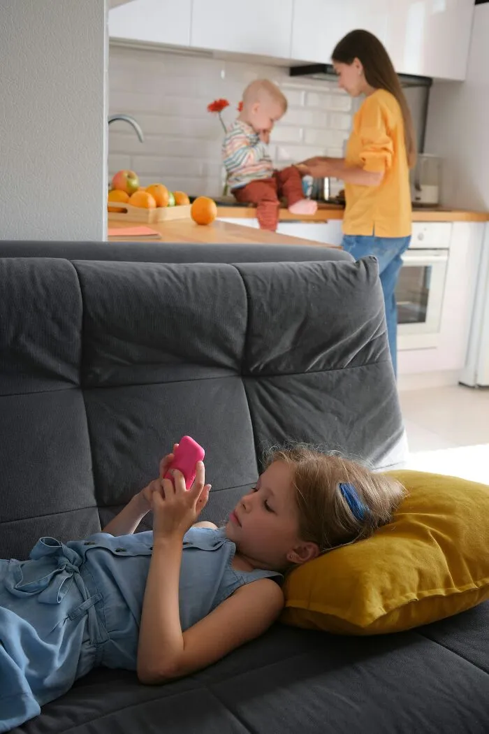 Child on a couch using a pink smartphone while a mother prepares food in the kitchen, illustrating normal modern behavior.