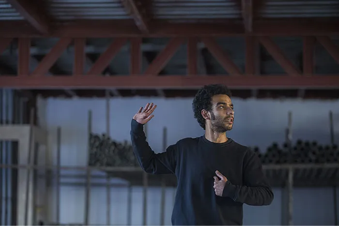 Person gesturing during a school presentation in a large room, wearing a dark sweater.