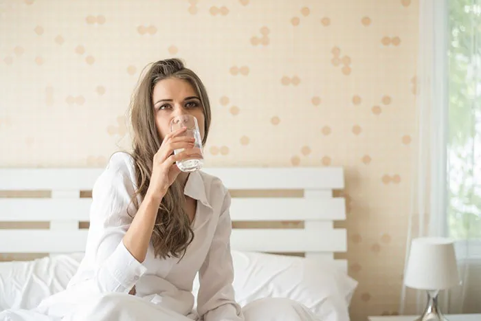 Woman in a white shirt drinking water in a bedroom, illustrating a life hack.