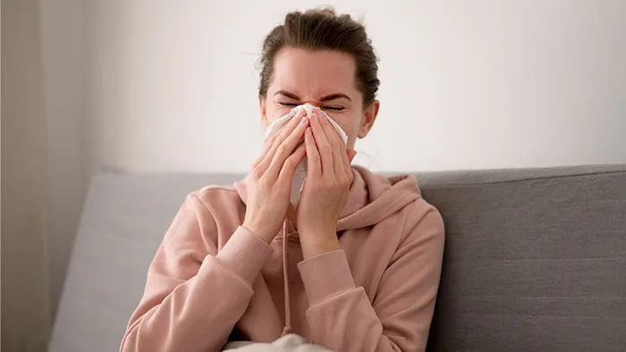 Woman sitting on a couch, sneezing into a tissue, demonstrating a crazy body life hack for dealing with colds.